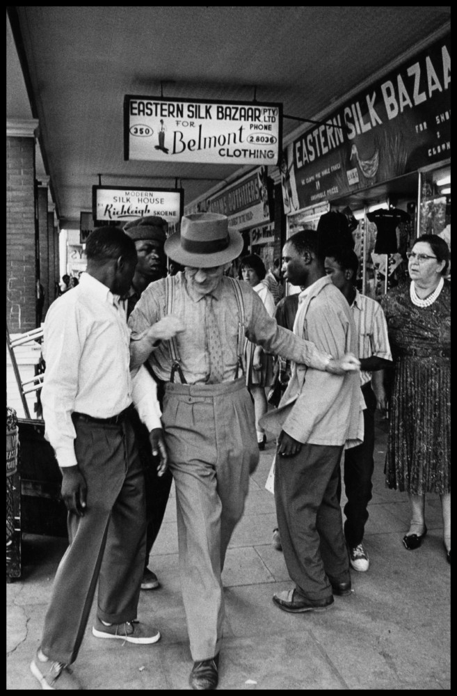Photo en Noir et Blanc montrant un homme blanc marchant sur un trottoir et repoussant un homme noir avec le coude (alors que d'autres lui piquent son portefeuille...)